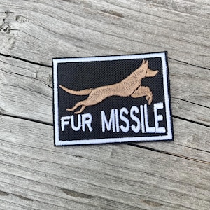 Pode incluir: Patch bordado com fundo preto e borda branca. O patch apresenta um cão marrom em pleno salto e as palavras "FUR MISSILE" em letras brancas.