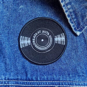 May include: Black and white embroidered patch of a vinyl record with the text "Greatest Hits" around the edge.