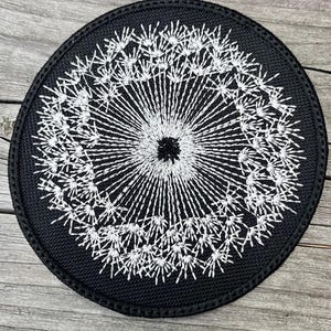 May include: A round, black patch with a white embroidered dandelion design. The dandelion is centered, with detailed stitching radiating outwards. The patch has a black border and is set against a wooden surface.