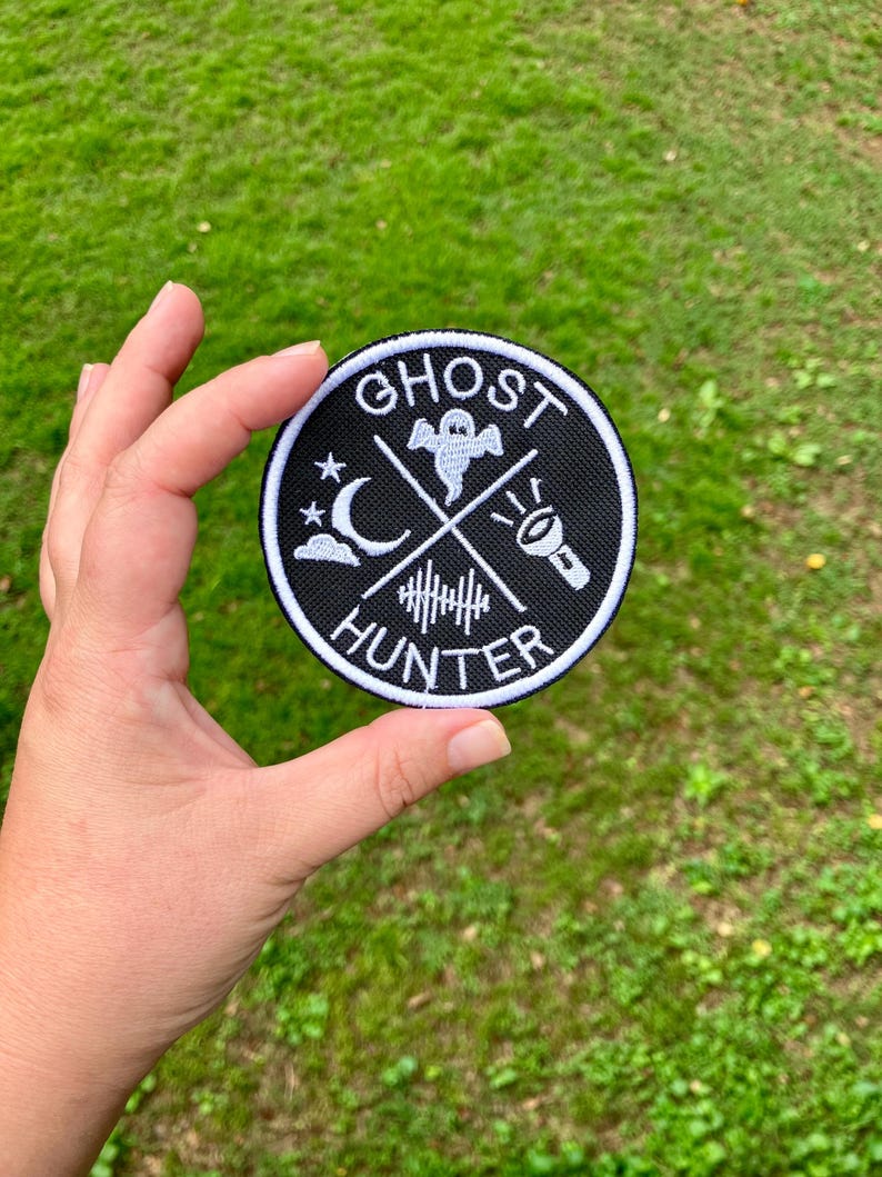 K&ouml;nnte beinhalten: Schwarz-wei&szlig; gestickter Aufn&auml;her mit dem Text "Ghost Hunter" in einem Kreis. Der Aufn&auml;her zeigt ein Gespenst, einen Halbmond, eine Taschenlampe und ein Kreuz.