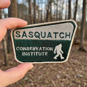 Puede incluir: Parche bordado verde y blanco con el texto "Sasquatch Conservation Institute" y una silueta blanca de un sasquatch caminando.
