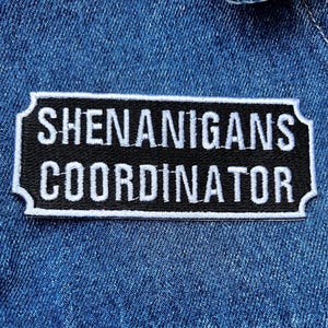 May include: A black and white embroidered patch with the words "SHENANIGANS COORDINATOR" in white capital letters. The patch has a black background and a white border, and is set against a blue denim background.