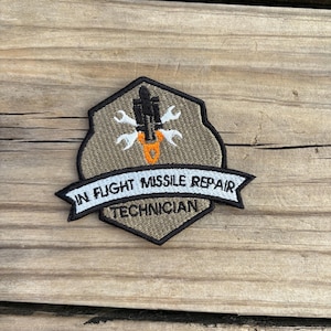 May include: Embroidered patch with a shield shape, featuring a missile and wrench design in white and orange on a tan background. The patch has the text "IN FLIGHT MISSILE REPAIR TECHNICIAN" in white on a black banner.