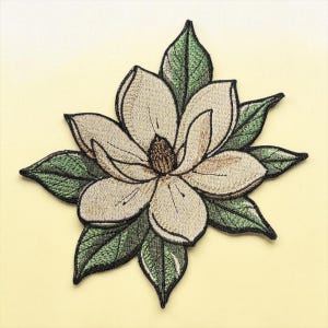 Op de afbeelding: Geborduurde magnolia bloem patch met crèmekleurige bloemblaadjes en groene bladeren. Het centrum van de bloem is donkerbruin en de bladeren hebben een gedetailleerd, getextureerd uiterlijk. De patch is geplaatst tegen een zachte gele achtergrond.