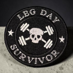 May include: A circular black embroidered patch featuring a white skull centered between two crossed barbells. The text LEG DAY is arched above the skull, and the word SURVIVOR is arched below. Two small white stars are positioned on the left and right sides.