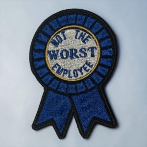May include: Embroidered blue ribbon patch with the text "NOT THE WORST EMPLOYEE" in white and gold. The patch has a black border and a blue ribbon design, suitable for jackets, bags, or other fabric items.