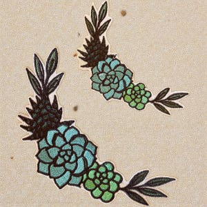 以下が含まれることがあります： 多肉植物のデザインが施された刺繍パッチが2つ。各パッチは、濃い緑色のパイナップルのような上部、明るい青緑色の多肉植物の花、濃い緑色の葉を持つ小さな緑色の多肉植物のつぼみを持っています。パッチはベージュ色の背景にあります。