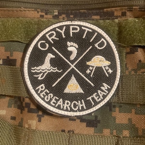 Peut inclure: Un écusson brodé noir et blanc avec le texte "Cryptid Research Team" en cercle. L'écusson représente une empreinte de pas, un canard, un OVNI et un triangle avec un œil à l'intérieur.