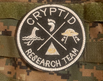 Puede incluir: Un parche bordado en blanco y negro con el texto "Cryptid Research Team" en un círculo. El parche presenta una huella, un pato, un OVNI y un triángulo con un ojo dentro.