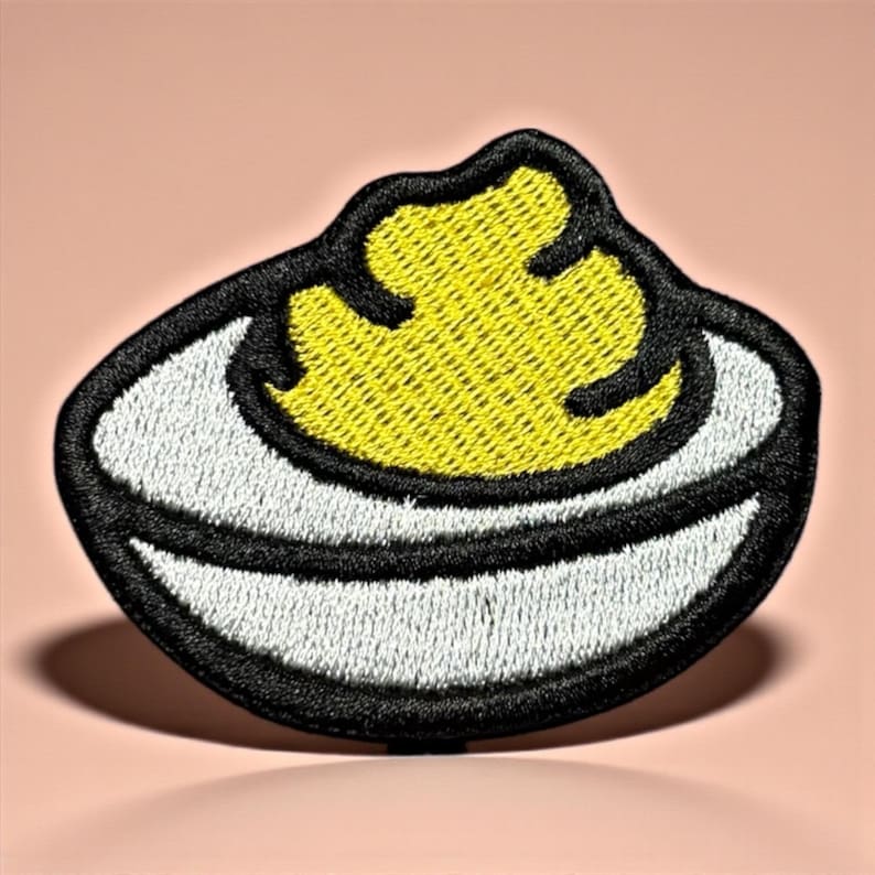 Puede incluir: Parche bordado con un dise&ntilde;o de huevo relleno. El parche tiene una clara de huevo blanca con un contorno negro y una yema amarilla. El parche est&aacute; sobre un fondo rosa.