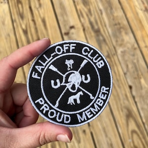May include: A round black and white patch with the text "FALL-OFF CLUB PROUD MEMBER" encircling a skull, crossed riding crops, a horseshoe, and a horse silhouette. The patch is held against a wooden background.
