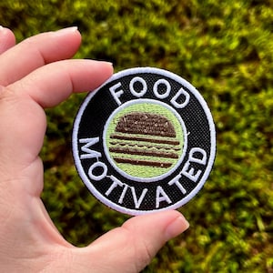Puede incluir: Un parche redondo con las palabras "FOOD MOTIVATED" en letras blancas alrededor de un gráfico de hamburguesa marrón y verde. El parche tiene un fondo negro con un borde blanco.