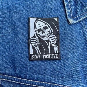 Puede incluir: Parche rectangular en blanco y negro con un esqueleto haciendo una señal de pulgar hacia arriba. El texto "STAY POSITIVE" está bordado en blanco debajo. El parche está cosido a una chaqueta vaquera azul.