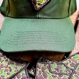Peut inclure: Casquette de baseball vert olive avec un écusson en forme de contour de l'État de la Caroline du Sud. L'écusson présente un motif camouflage vert, marron et beige. Plusieurs écussons assortis sont visibles.