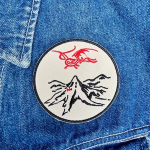 May include: Close-up of a round embroidered patch with a black border. The patch features a red dragon and a black mountain range design on a cream background. The patch is sewn onto a blue denim jacket.