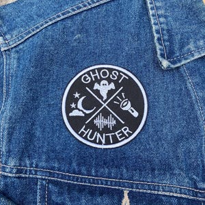 K&ouml;nnte beinhalten: Ein gestickter Patch in Schwarzwei&szlig; mit dem Text "Ghost Hunter" in einem Kreis. Der Patch zeigt ein Gespenst, einen zunehmenden Mond, eine Taschenlampe und eine Schallwelle.