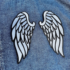 Op de afbeelding: Twee geborduurde vleugelpatches in lichtblauw en wit, omrand met zwart, worden getoond op een spijkerjasje. De patches zijn symmetrisch, met gedetailleerde veermotieven.