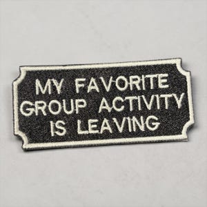 May include: A rectangular black patch with white embroidered text that reads "MY FAVORITE GROUP ACTIVITY IS LEAVING." The patch has a white border and a decorative shape.