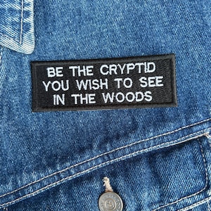 May include: A close-up of a blue denim jacket with a black rectangular patch. The patch features white text that reads, "BE THE CRYPTID YOU WISH TO SEE IN THE WOODS." The jacket has a button and white stitching.