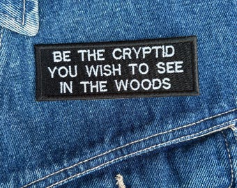 May include: A close-up of a blue denim jacket with a black rectangular patch. The patch features white text that reads, "BE THE CRYPTID YOU WISH TO SEE IN THE WOODS." The jacket has a button and white stitching.