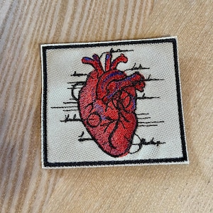 Puede incluir: Un parche bordado beige con borde negro que presenta un corazón anatómico rojo con venas azules. El corazón está rodeado de líneas negras.
