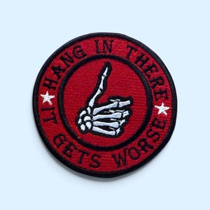 Puede incluir: Un parche bordado circular con fondo rojo y borde negro. En el centro hay una mano de esqueleto blanca haciendo un gesto de pulgar hacia arriba. El texto "HANG IN THERE IT GETS WORSE" está bordado en negro alrededor del borde con dos estrellas.