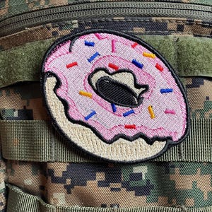 May include: A pink and white embroidered donut patch with colorful sprinkles. The patch is attached to a green camouflage backpack.