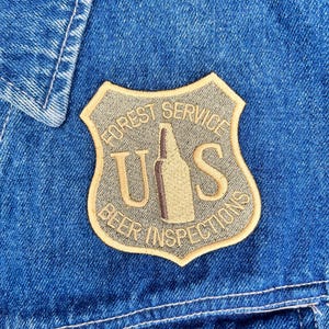 May include: Close-up of a blue denim jacket with a Forest Service Beer Inspections patch. The patch is shield-shaped with the text "FOREST SERVICE" at the top, "U S" in the center with a beer bottle graphic, and "BEER INSPECTIONS" at the bottom.