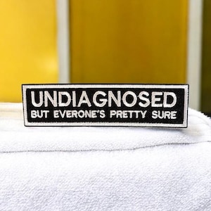 May include: A rectangular black and white patch with the text "UNDIAGNOSED BUT EVERONE'S PRETTY SURE". The patch is on a white, textured surface, with a yellow background.
