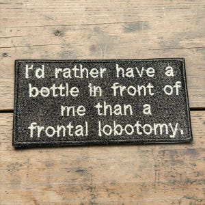 May include: A rectangular, black embroidered patch with white text that reads, "I'd rather have a bottle in front of me than a frontal lobotomy." The patch is set against a wooden background.