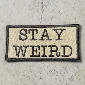 May include: A rectangular patch with the words "STAY WEIRD" embroidered in black on a tan background. The patch has a black border and is likely designed to be sewn or ironed onto clothing or accessories.