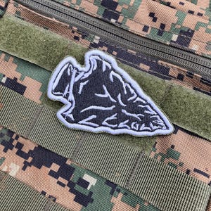 Puede incluir: Un parche de punta de flecha negro y blanco con un contorno blanco. El parche está unido a una tela de camuflaje verde y marrón.