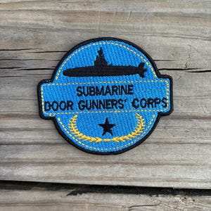 May include: Embroidered patch featuring a black submarine silhouette on a blue background. The text "SUBMARINE DOOR GUNNERS' CORPS" is displayed above a black star and a gold wreath. The patch has a black border.