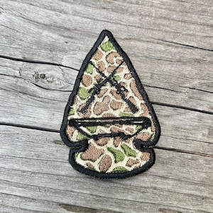 Könnte beinhalten: Gestickter Aufnäher in Pfeilspitzenform. Das Tarnmuster ist in Braun-, Grün- und Beigetönen gehalten. Zwei schwarze Gewehre kreuzen sich auf dem Muster. Der Aufnäher ist schwarz umrandet.