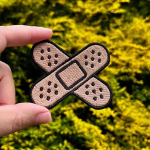 May include: Embroidered patch in the shape of two overlapping bandages. The bandages are tan with black stitching and small black dots. The patch has a black border and is held against a blurred green and yellow background.