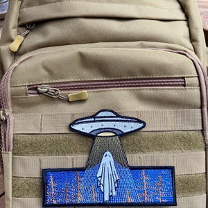 May include: A tan tactical backpack with a black and blue embroidered patch depicting a UFO hovering over a ghost in a forest.