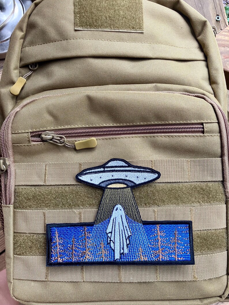 May include: A tan tactical backpack with a black and blue embroidered patch depicting a UFO hovering over a ghost in a forest.