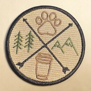 Può includere: Toppa ricamata con un design circolare con un'impronta di zampa, montagne, alberi e una tazza di caffè. Frecce nere si incrociano al centro. La toppa ha uno sfondo beige con un bordo nero.