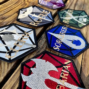May include: Embroidered hexagon-shaped patches with different designs and text. The patches feature the words "ROGUE", "RANGER", "SORCERER", and "BARBARIAN". The patches are in various colours and are displayed on a wooden surface.