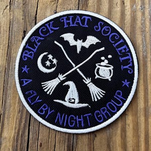 Op de afbeelding: Ronde zwarte patch met wit en paars geborduurde tekst en afbeeldingen. De tekst luidt "BLACK HAT SOCIETY" en "A FLY BY NIGHT GROUP". Afbeeldingen zijn onder meer een vleermuis, maan, heksenhoed, gekruiste bezems en een ketel.