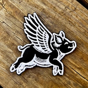 Puede incluir: Parche en blanco y negro que representa un cerdo volador con alas blancas. El parche está bordado con un diseño detallado y tiene un borde negro. Se encuentra sobre una superficie de madera.