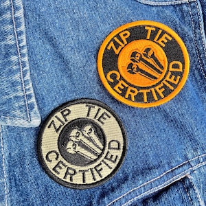 May include: Two circular patches with the text "ZIP TIE CERTIFIED" embroidered in a circular pattern. One patch is black and orange, the other is black and tan. The patches are on a blue denim jacket.