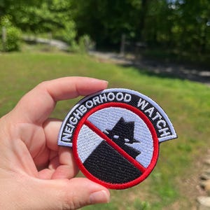 May include: Embroidered patch with the words "NEIGHBORHOOD WATCH" in white on a black and white background. The patch features a red circle with a crossed-out silhouette of a person wearing a hat.