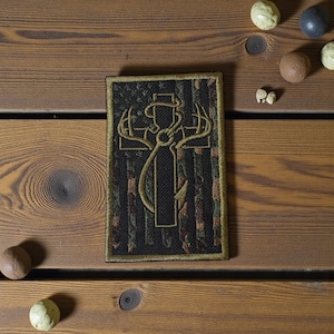 May include: Embroidered patch with a gold cross design, deer antlers, and a cowboy hat. The patch has a black camouflage background and a gold border. The rectangular patch is displayed on a wooden surface with various small, round objects.