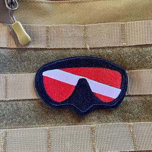 Peut inclure: Un patch de masque de plongée sous-marine rouge, blanc et noir avec un dos en velcro. Le patch est fixé à un panneau molle beige.