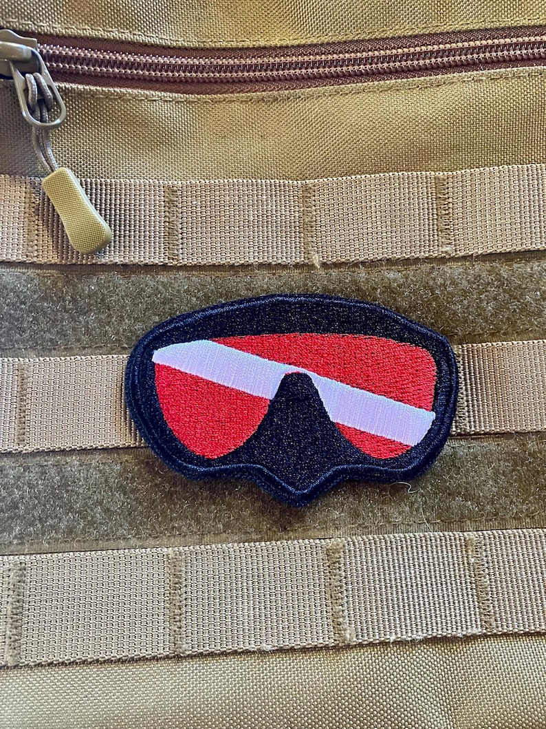 May include: A red, white, and black scuba diving mask patch with a velcro backing. The patch is attached to a tan molle panel.