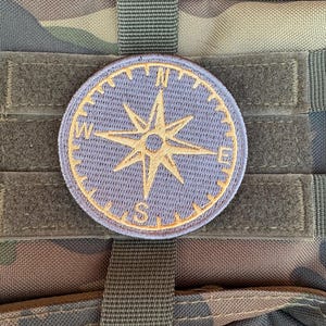 May include: A round, light blue compass patch with a gold-colored compass rose design. The patch is attached to a camouflage-patterned fabric with hook-and-loop fasteners and a dark green strap.
