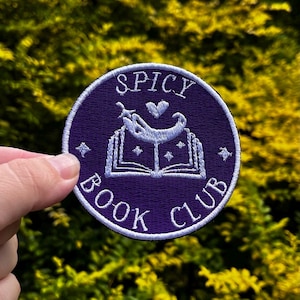 May include: A round, purple patch with white embroidered text and design. The text reads "SPICY BOOK CLUB" around the edge. The design features an open book with a chili pepper and heart above it. The patch is held by a hand.