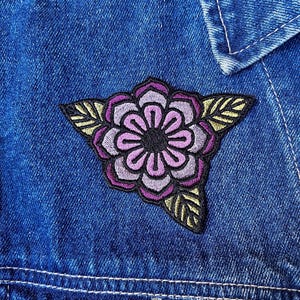 Könnte beinhalten: Nahaufnahme einer blauen Jeansjacke mit einem Blumenaufnäher. Der Aufnäher zeigt eine lila und rosa Blume mit schwarzen und gelben Blättern. Die Jacke hat sichtbare Nähte und einen Knopfverschluss.