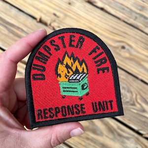 May include: Embroidered patch with the words "DUMPSTER FIRE" in a curved arch over "RESPONSE UNIT". The patch features a green dumpster on fire with orange and yellow flames, all on a red background with a black border.